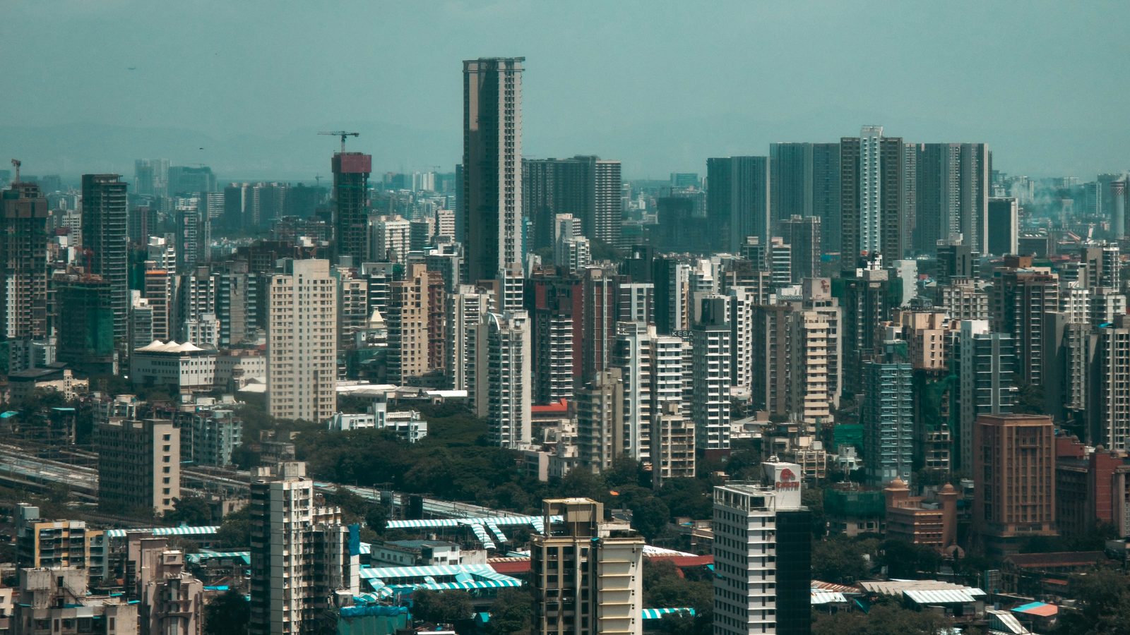 Dense high-rise Dadar seen from above, the railway tracks and striped platform canopies of Dadar station across the foreground, towers stretching to a hazy horizon