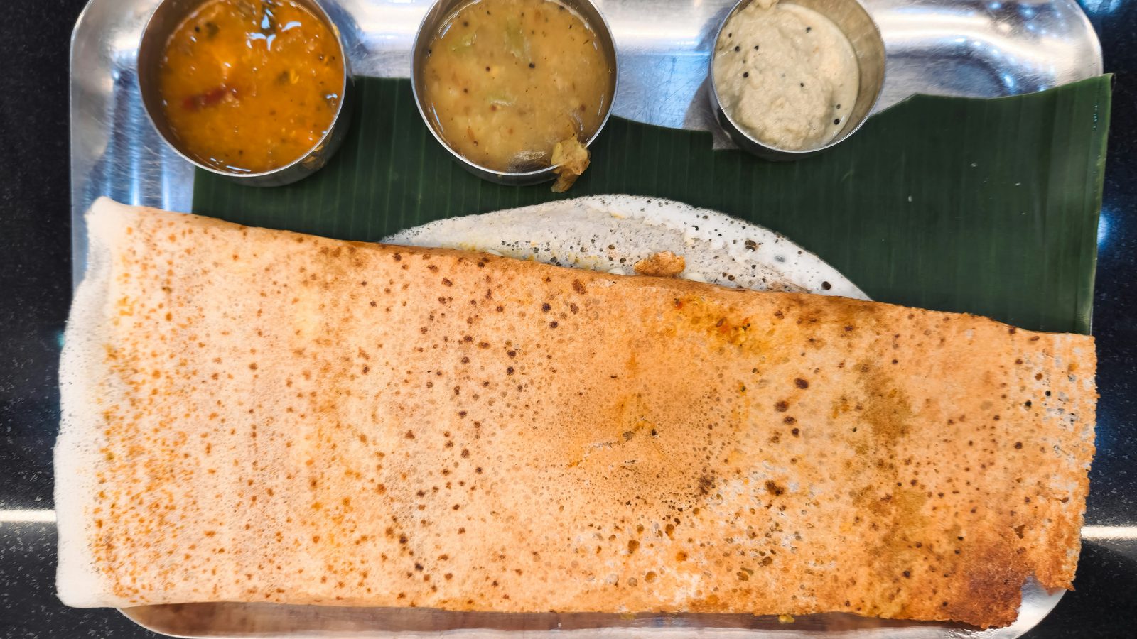 A dosa on a banana leaf and steel tray, with three chutneys