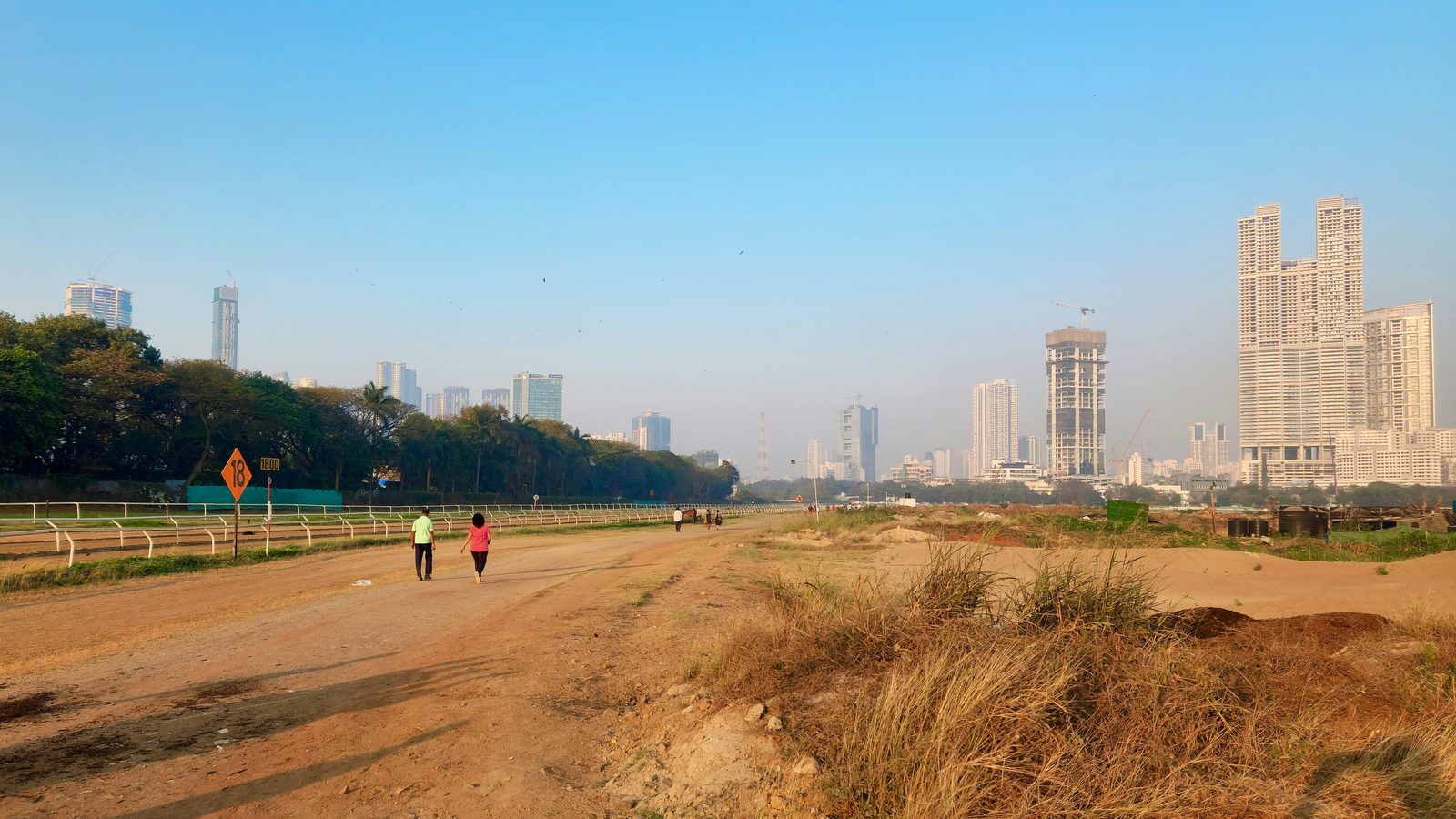 Open ground in Mumbai early in the morning, two walkers small on a dirt track, a treeline and the city skyline along the horizon