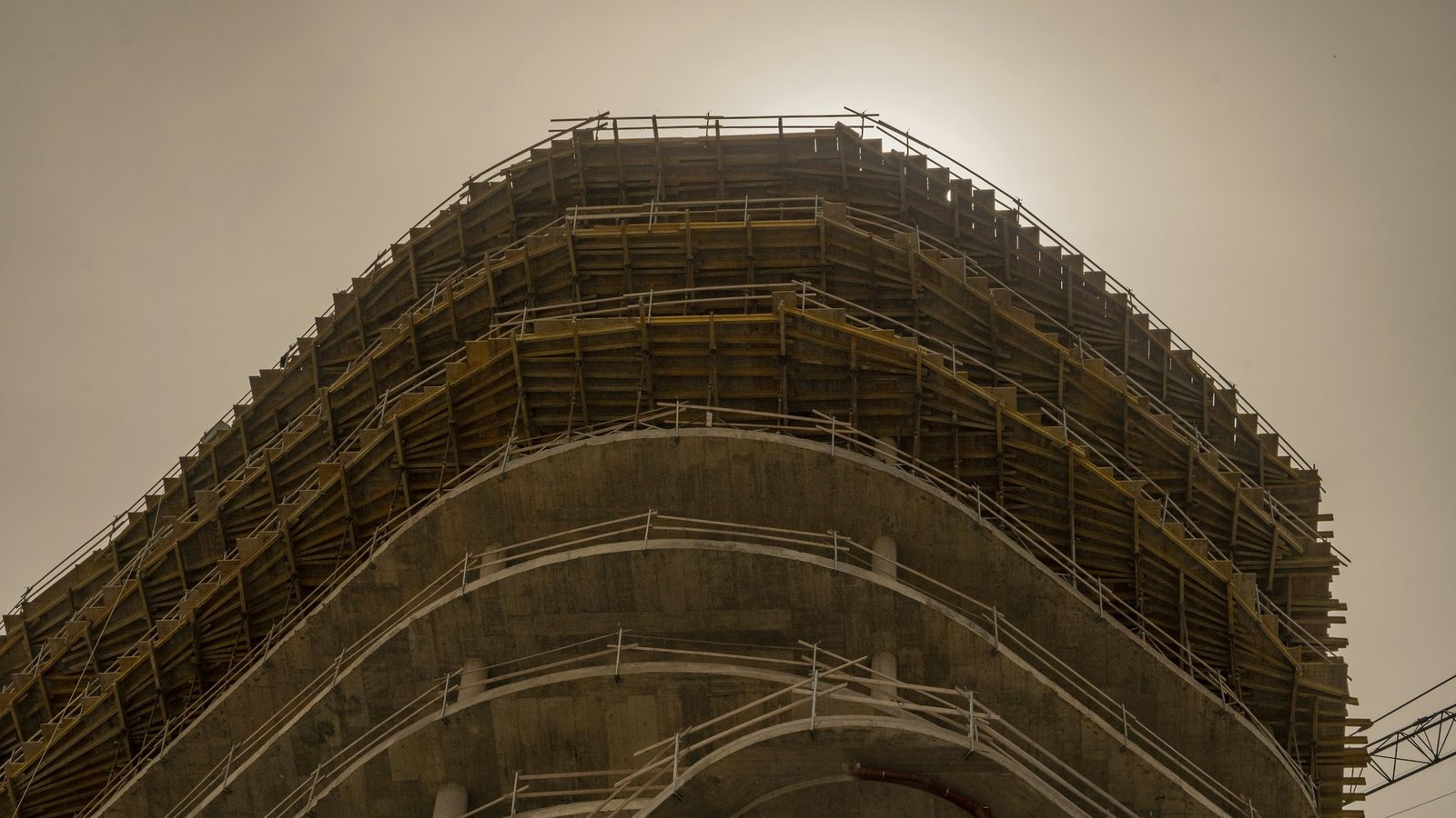 A curved high-rise under construction seen from below, bare concrete floor plates and railings stacked against a pale sky