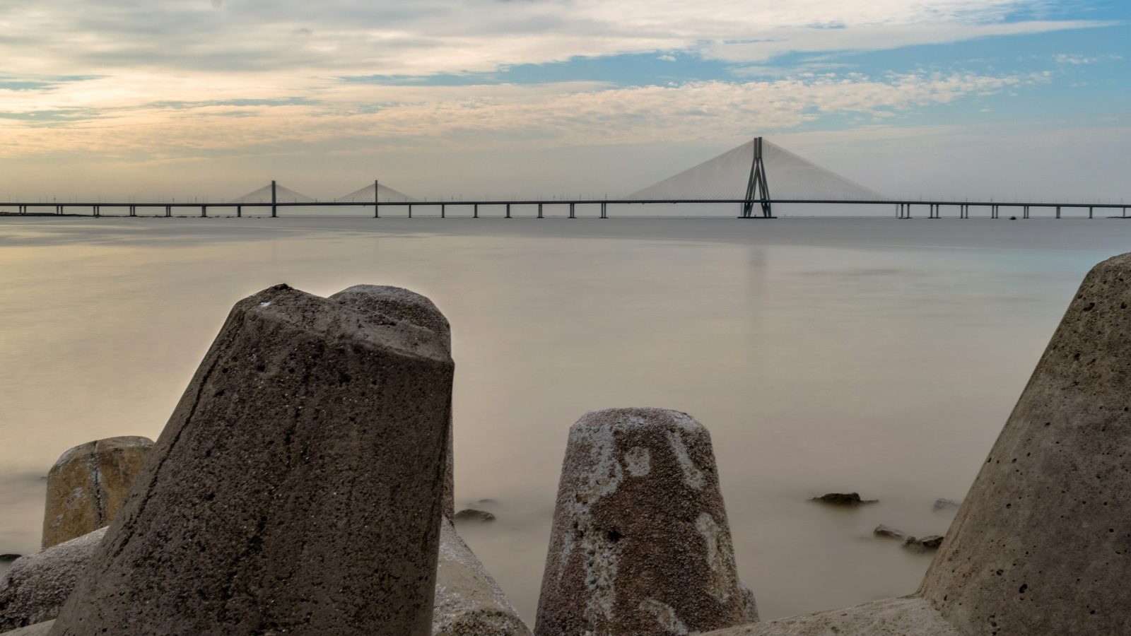 Concrete tetrapods on a Mumbai sea wall in the foreground, the Bandra-Worli Sea Link across calm water at golden hour